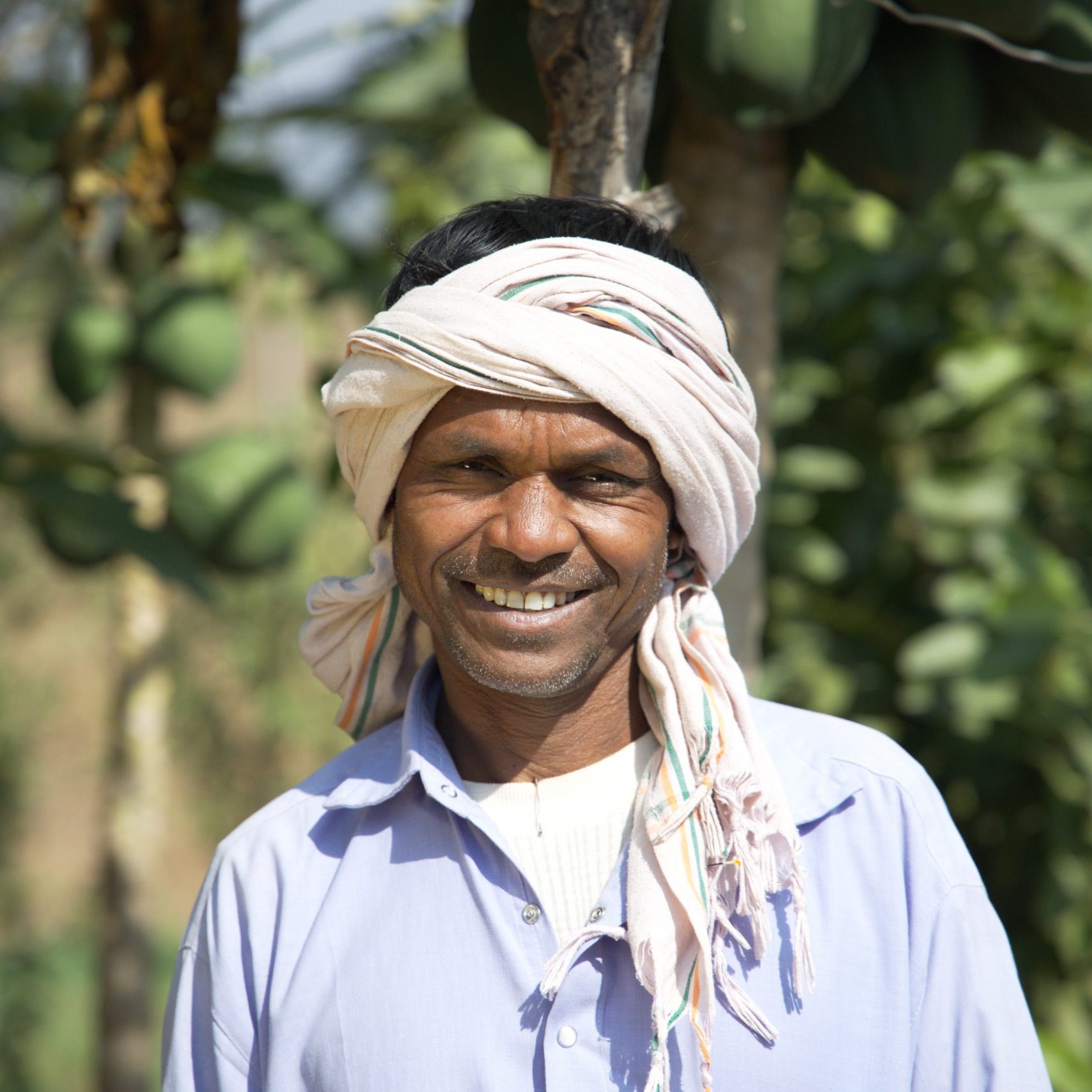 A man with a white head turban smiles at the camera, trees behind him.