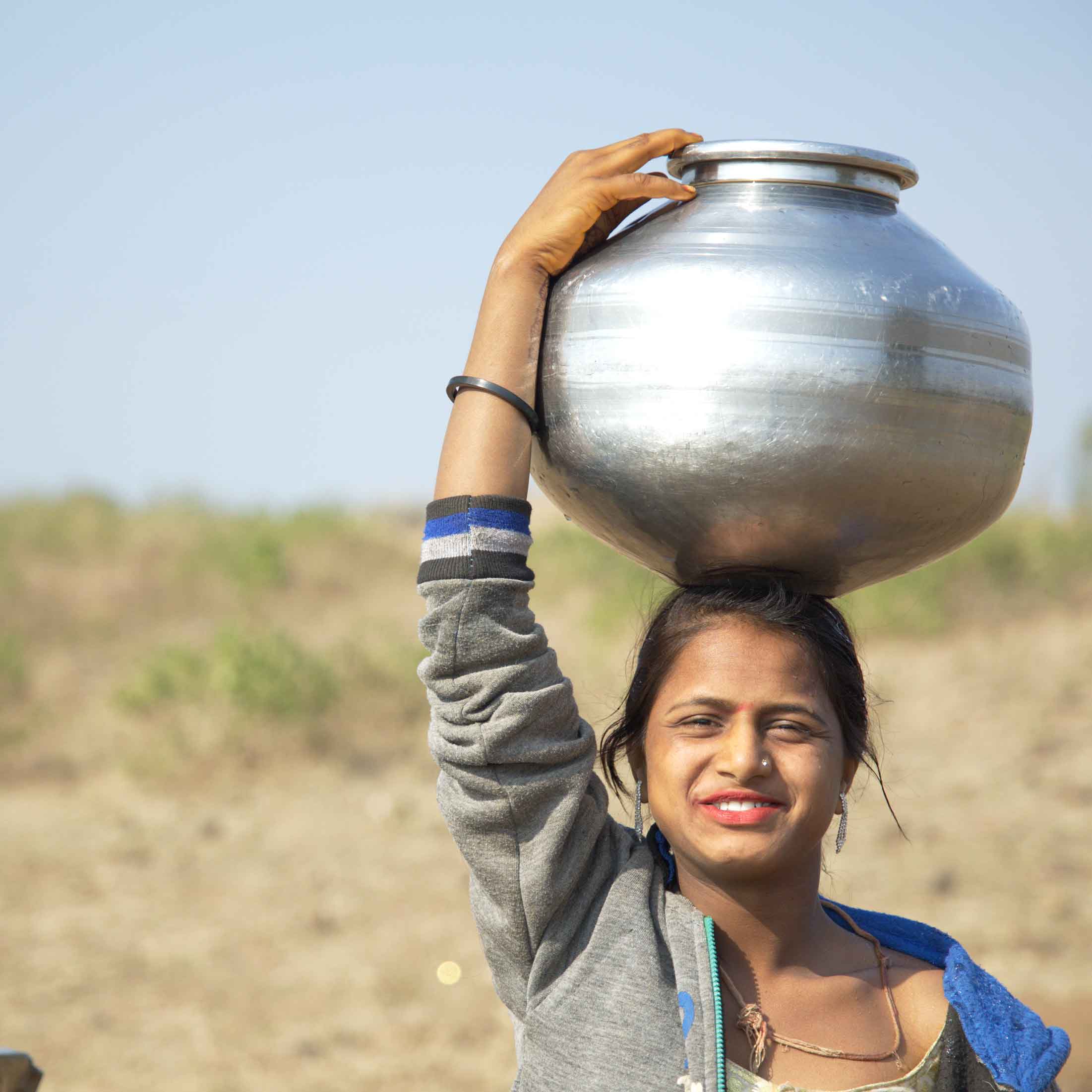 A lady holds a large silver pot on her head and stands in a barren field. 