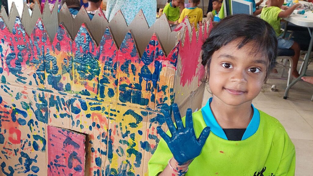 A small girl stands in front of a multi-coloured finger paint artwork, with a blue paint covered hand