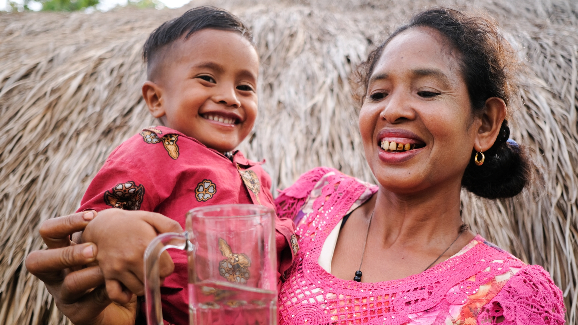 A woman and her child hold a glass of water and smile in front of their hut