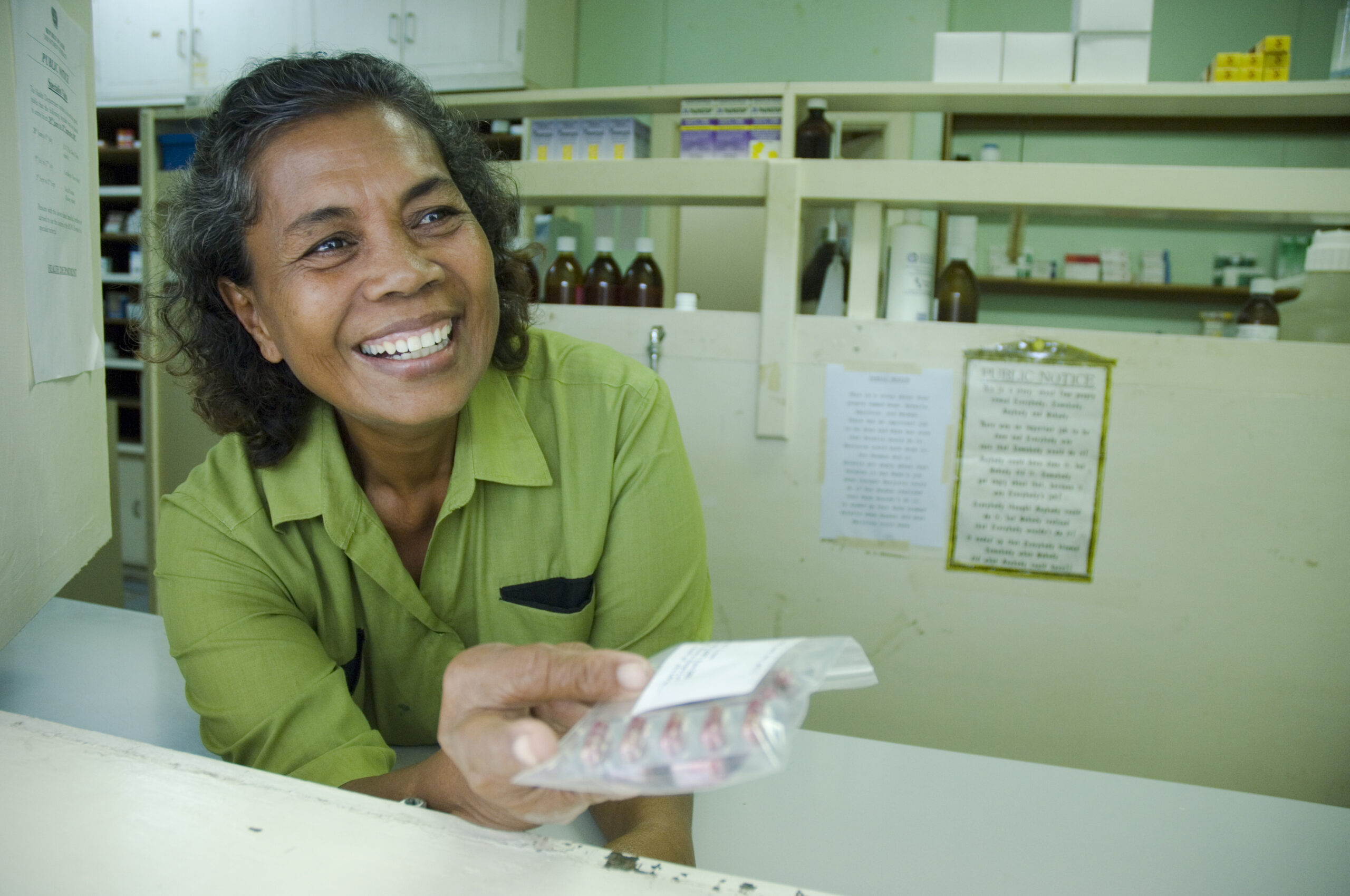 A smiling woman in a green shirt hands out medication at a pharmacy.