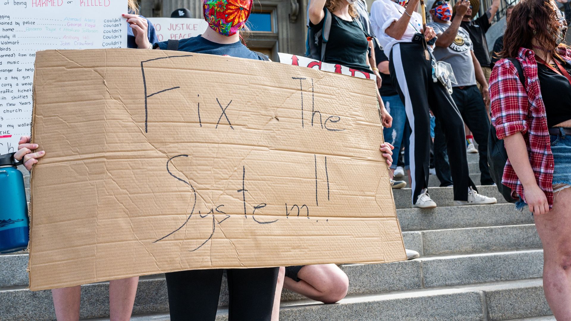A protestor with a floral mask holds up a cardboard sign that reads 'fix the system'